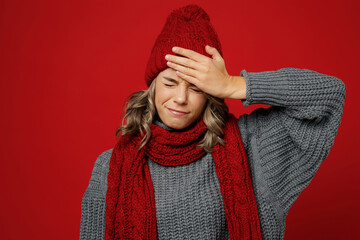 Wall Mural - Young tired exhausted sad woman wear gray sweater scarf hat put hand on forehead head suffer isolated on plain red background studio. Healthy lifestyle ill sick disease treatment cold season concept.