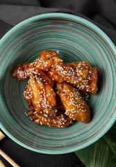 Poster - Roasted chicken wings in teriyaki sauce with sesame on ceramic bowl. Buffalo wings in asian style on black concrete background. Aesthetic composition with fried chicken wings, chopsticks and leaf.