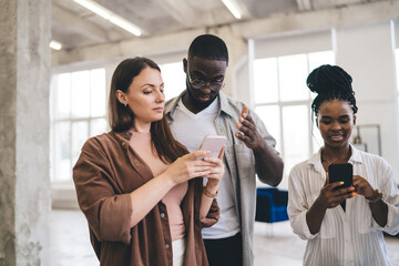 Sticker - Multiethnic coworkers standing in modern office discussing work while browsing smartphones