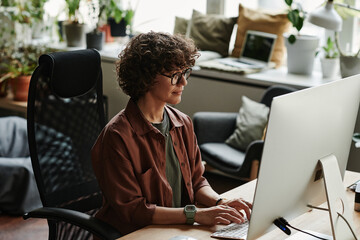 Young serious businesswoman typing on keyboard in front of computer monitor while working over new website in openspace office