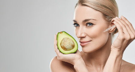 Wall Mural - Caucasian woman applying avocado oil on her face with pipette dropper for nourishing and moisturizing skin and holds half of avocado, high quality. Facial skin care with avocado oil