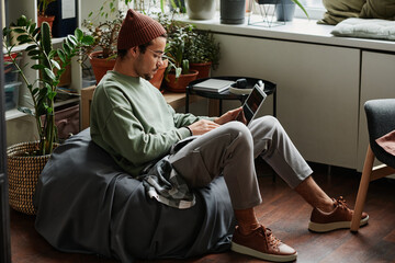 Leinwandbilder - Young serious webdesigner with laptop concentrating on network while sitting in comfortable armchair in office with many green plants
