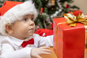 Wall Mural - Little boy with a Santa Claus hat looks at the Christmas gift on a table