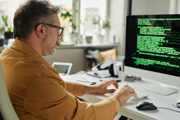Leinwandbilder - Side view of mature serious male IT engineer typing on computer keyboard while sitting in front of screen with coded data