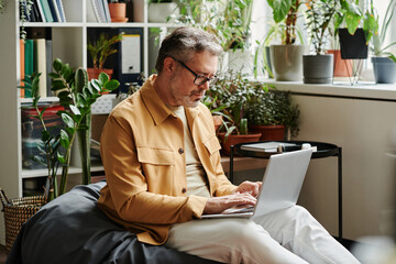 Leinwandbilder - Serious mature businessman in casualwear and eyeglasses with laptop on knees networking in office while sitting on soft bag chair