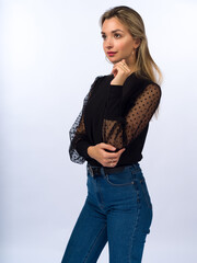 A beautiful, young, slender girl with long blonde hair on a white background in blue jeans and a stylish black blouse with transparent sleeves. Close-up