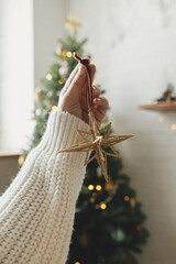 Wall Mural - Hand holding stylish christmas golden star on background of christmas tree in lights in festive boho room. Decorating christmas tree with golden ornament. Atmospheric time