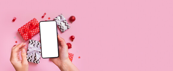 Online Christmas shopping concept. Female hand with phone mock up and Christmas presents flat lay on a pink background