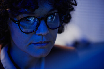 Poster - Selective focus of face of serious businesswoman in eyeglasses sitting in front of computer late at night in office and working overtime
