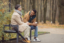 Friends, A Senior And A Young Man Sit In The Park On A Bench And Talk In The Autumn Park.