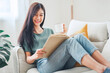 © oatawa - Young asian woman sitting on modern sofa in front of window relaxing in her living room reading book and drinking coffee or tea at home