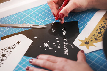 christmas graphic cut from black adhesive foil is finished by female hands with painted nails. weeding tool, scalpel and additional stickers on worktop. closeup. holiday gift and advertisement concept