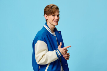 a handsome friendly attractive red-haired young man student stands with a red backpack and a fashionable jacket on a light blue background and shows hand gestures. Horizontal photo with empty space