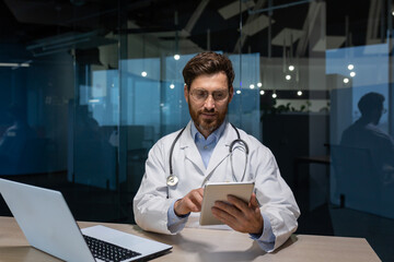 mature doctor in a medical gown is working inside clinic office, doctor is holding a tablet computer in his hands for online consultation of patients, working with a laptop.