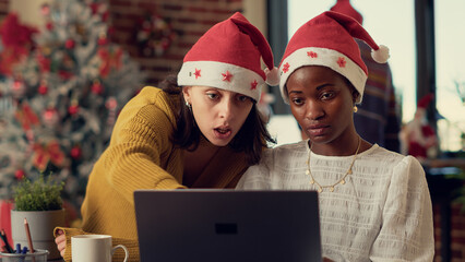 Multiethnic team of coworkers planning report on laptop, using research data in office with christmas tree and ornaments. Diverse people with santa hats celebrating xmas winter holiday.