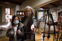 Curious Tween Schoolgirl And Friendly Elderly Female Tutor In Face Masks Viewing Vintage Bicycle In Museum Of History Of Technology With Interest. Knowledge Concept. New Normal In Coronavirus Pandemic