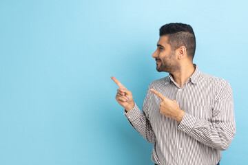 Smiling bearded businessman pointing aside, showing blank copy space for idea presentation, commercial text, looking away, wearing striped shirt. Indoor studio shot isolated on blue background.