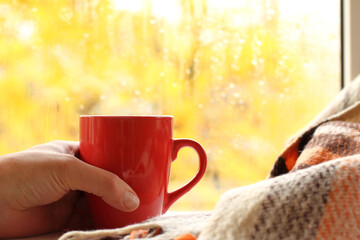 Wall Mural - red mug in hand against the background of a window with an autumn landscape. warm morning cozy