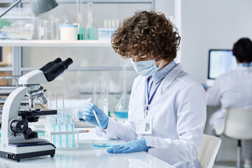 young biochemist in protective workwear bending over desk while mixing two liquid substances in petr