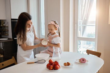 An image of a young woman and her little cute daughter are cooking in the kitchen. Have fun together, interfere with flour. Mom teaches child to cook