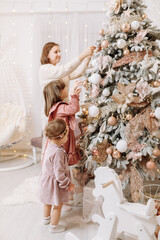 Pretty smiling woman with adorable two daughters decorate Christmas tree at home. Joyful little girls with mother spend winter holidays, enjoy happy moments, childhood and motherhood concept.