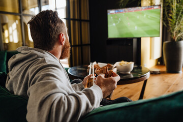 Canvas Print - Excited white man holding beer and pizza while watching football match on TV