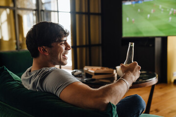 Wall Mural - Young white man watching football match and drinking beer in front of the TV screen