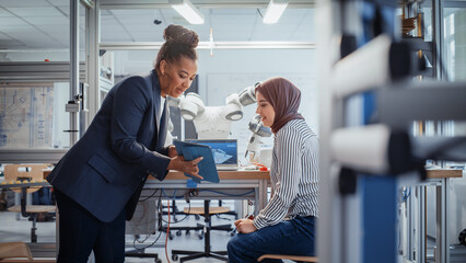 Black Senior Engineer Discussing Robotic Arm with Promising Arabic Female Computer Scientist While Manipulating it with Laptop and Tablet. Manufacturing Facility that Creates High Tech Products.