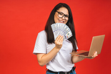 Canvas Print - Happy winner! Pretty young business woman in casual holding laptop in the office, isolated over red background. Working with computer, holding money bills.