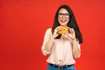 Wall Mural - Portrait of young beautiful hungry woman eating burger. Isolated portrait of student with fast food over red background. Diet concept.