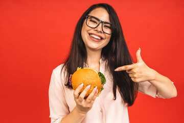 Wall Mural - Portrait of young beautiful hungry woman eating burger. Isolated portrait of student with fast food over red background. Diet concept.