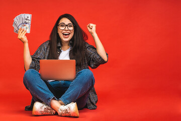 Canvas Print - Business concept. Portrait of happy brunette woman in casual sitting on floor in lotus pose and holding laptop isolated over red background. Holding money bills.