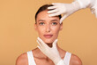 Cosmetic surgery concept. Doctor examining young caucasian woman's face over beige studio background