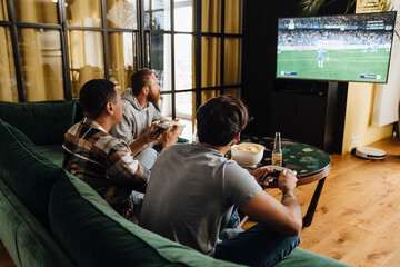 Wall Mural - Young men playing football video game with gamepads at home