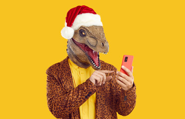 Wall Mural - Funny excited dinosaur man in Christmas hat using mobile phone. Male fashion model wearing weird wacky bizarre dino lizard mask, leopard jacket and Xmas cap holding smartphone and sending text message