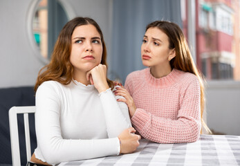 Upset young woman sitting at home table with disgruntled woman behind