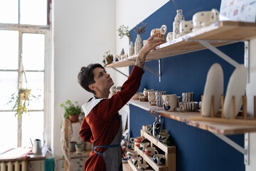 Wall Mural - Small business female owner putting plate at right place in pottery studio. Self employed girl pottery studio owner tidies up the shelves with handcraft ceramics at her cozy workplace