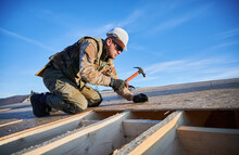 Carpenter Hammering Nail Into OSB Panel On The Roof Top Of Future Cottage On Blue Sky Background. Man Worker Building Wooden Frame House. Carpentry And Construction Concept.