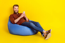 Full Size Photo Of Positive Young Man Sit Comfy Bag Hold Use Telephone Chatting Isolated On Yellow Color Background
