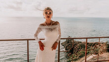 Romantic Bride, Blond Girl In White Wedding Dress With Open Shoulders Posing On Open Terrace With Backdrop Of The Sea And Rocks. Stylish Young Woman Standing On Terrace And Looking On Ocean At Sunset