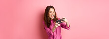 Technology Concept. Carefree Young Woman Tilt Body, Holding Smartphone Horizontally, Playing Video Game On Phone, Standing Against Pink Background