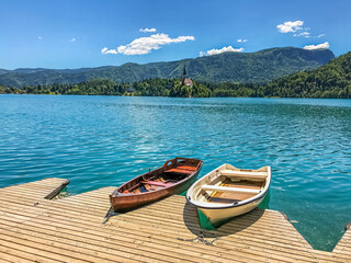 Two wooden boats by the wooden pier against the backdrop of a chapel and wooded hills on Lake Bled in Slovenia on a sunny day.