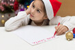 Cute little girl in Santa hat is writing a letter to Santa Claus on Christmas. Shallow depth of field, focus on the hand
