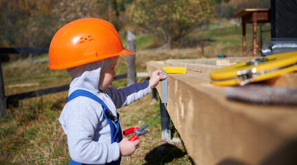 Wall Mural - Boy toddler playing as builder on construction site. Child carpenter in orange helmet and blue overalls learning to build wooden frame house outdoor on sunny day. Carpentry and workshop concept.