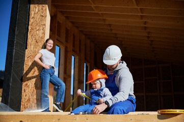 Wall Mural - Father, mother and son building wooden frame house in the Scandinavian style barnhouse. Toddler boy helping his daddy, while woman looking for them on construction site. Carpentry, family concept.