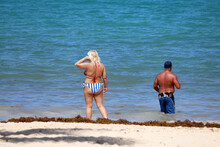 Beach Holiday And Vacation, Couple Going To Swim In The Sea. Blonde Woman With Cellulite Wearing Bikini And Tanned Man In Swimming Trunks Walk On Sand With Algae
