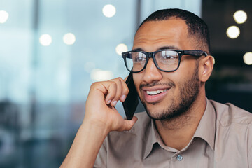 Close-up photo of happy and smiling african american businessman in glasses talking cheerfully on phone with customers, male salesman working inside modern office building.