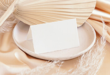 Wall Mural - Folded card near dried palm leaf and beige silky fabric close up, greeting or wedding mockup