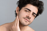 Fototapeta  - A young caucasian man with stubble beard is moisturizing face and skin with cosmetic oil while doing a facial massage, looking into the camera