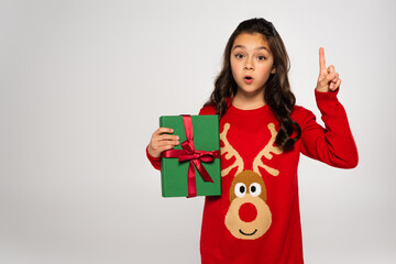 Wall Mural - surprised girl in red sweater holding wrapped Christmas present and pointing with finger isolated on grey